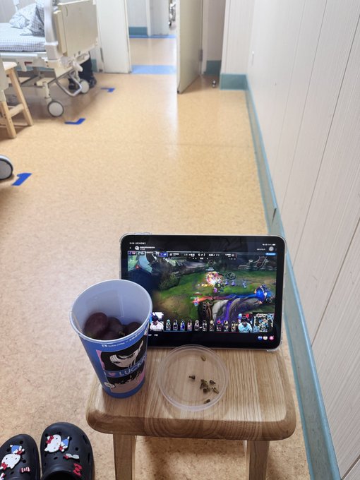 A tablet displaying a League of Legends match sits on a wooden stool in what appears to be a hospital hallway, next to a blue cup and a small lid with seeds.