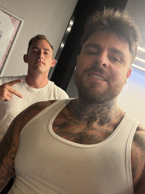 Two men pose for a selfie in a hotel room—the bearded man in front wears a white tank top and sports prominent chest and neck tattoos, including visible script that reads "Believe" and "trust your hustle," while a younger man in a white t-shirt makes a hand sign behind him. The image connects to the post by showing the team gearing up for setup day at the XRP booth for RareEvo.