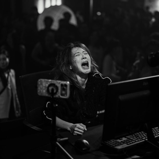 A black and white photo of a young woman screaming in apparent frustration or excitement while playing a video game at a computer, with a phone mounted on a tripod in the foreground.