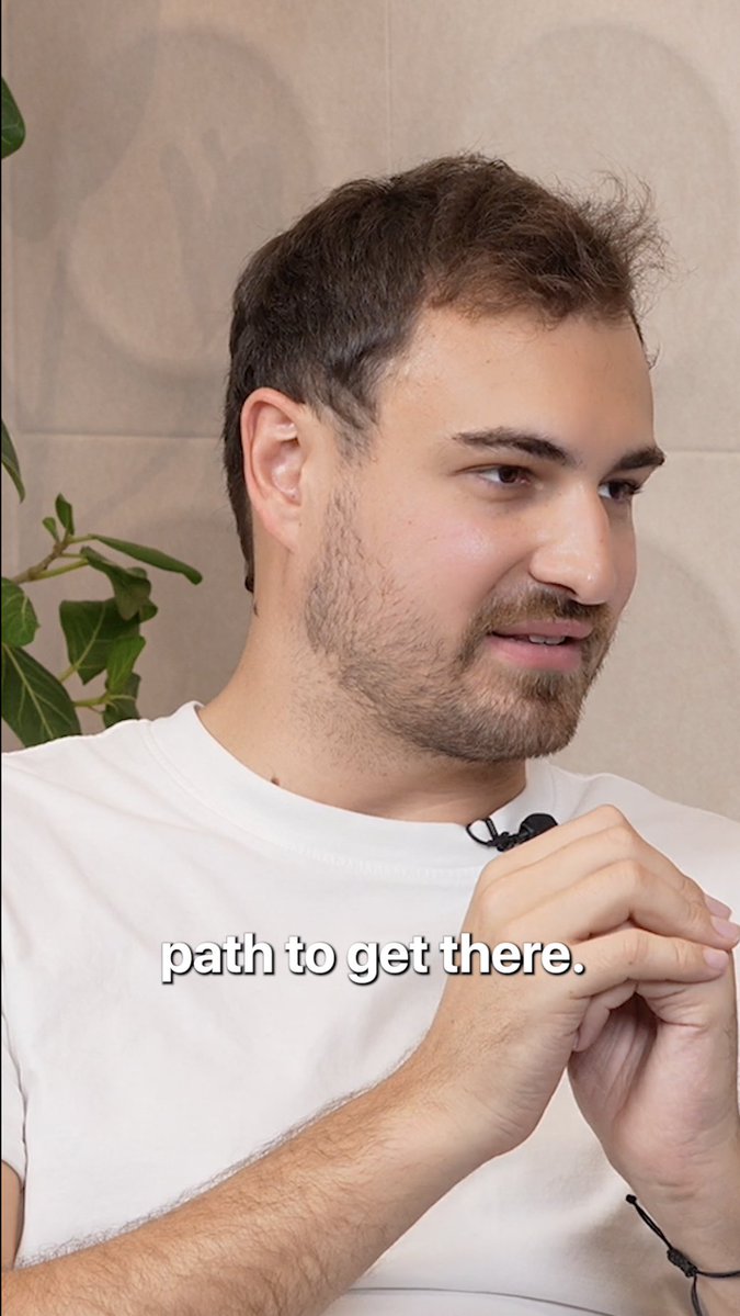 A man with brown hair and a light beard, wearing a white t-shirt with a lapel microphone, looks thoughtfully off-camera with his hands clasped. The on-screen text reads "path to get there." The image appears to be from a video interview about evaluating founders, capturing a moment of focused, contemplative discussion.