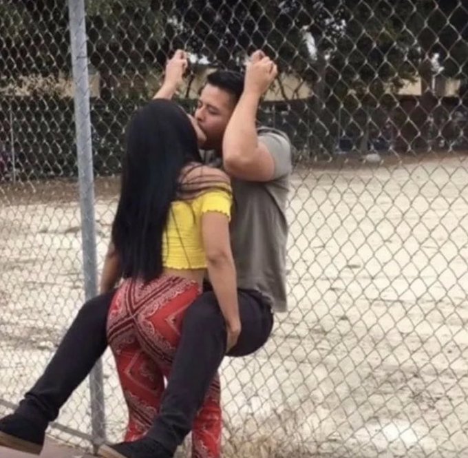 A couple kissing through a chain-link fence, with the woman in a yellow top and red patterned pants holding up the man in a grey shirt.