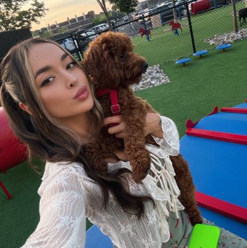 A woman with long brown hair poses for a selfie while holding a small brown poodle, taken outdoors at what appears to be a dog park with agility equipment.