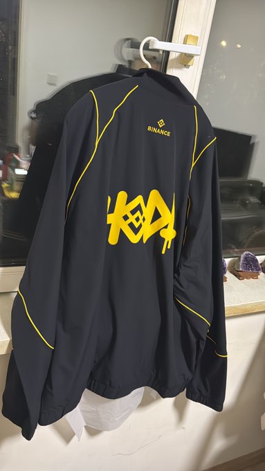 A black and yellow jacket with the Binance logo and a graffiti-style "9" design hangs on a hanger, commemorating Binance's 9th anniversary celebration.