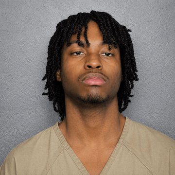 A young man with shoulder-length twisted locs poses for what appears to be a mugshot, wearing a beige V-neck garment against a plain gray wall. The image likely depicts the hacker referenced in the accompanying post, who was arrested by the FBI for allegedly spreading crypto-stealing malware disguised as video games on Steam.