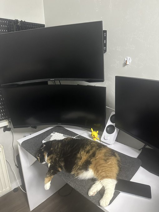 A calico cat sleeping on a felt desk mat in front of a triple-monitor computer setup, with a small Pikachu figurine placed between the screens.