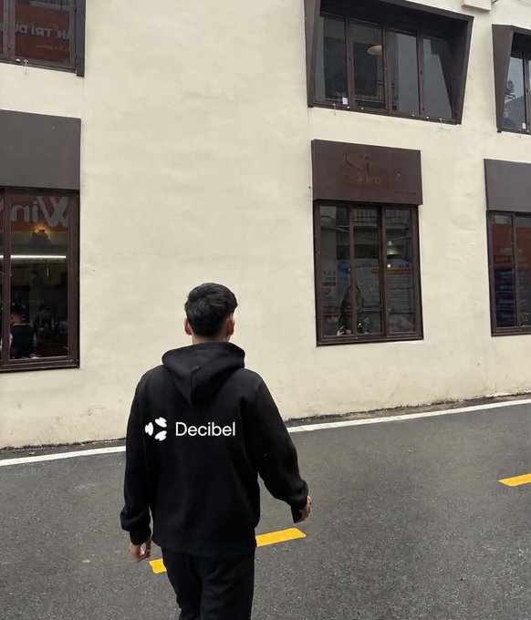 A man wearing a black "Decibel" hoodie walks down a narrow street, his back to the camera as he moves toward a cream-colored building with dark-framed windows. The image embodies the "gm suffer now, live in comfort later" mindset—capturing a quiet moment of daily grind, where the wearer pushes forward in branded comfort toward future ease.