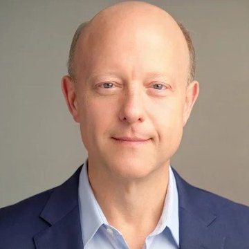 A professional headshot of a bald man in a blue suit jacket and light blue shirt smiling against a gray background.