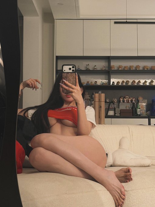 A woman in a red and white top takes a mirror selfie on a beige couch with shelves of collectible figurines in the background. The image doesn't directly relate to the post's discussion about BSC (Binance Smart Chain) and the Two Sages' tweets.