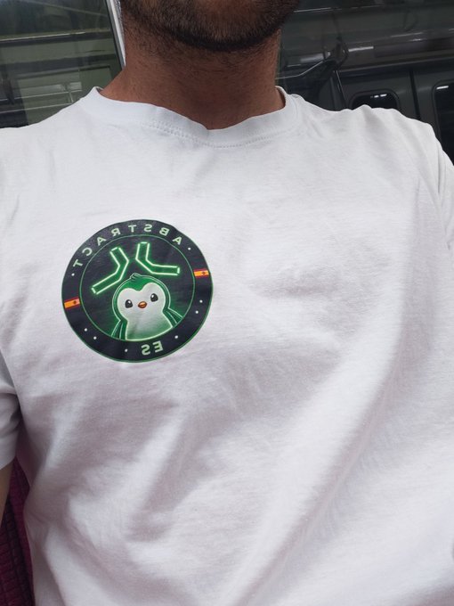 A white t-shirt featuring a circular green patch with a penguin character and the text 'TARTZBA' and '23'.
