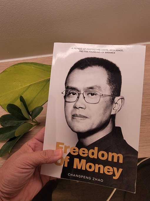 A hand holding up a book titled 'Freedom of Money' by Changpeng Zhao, subtitled 'A MEMOIR OF PROTECTING USERS, RESILIENCE, AND THE FOUNDING OF BINANCE,' with a green plant and wooden table in the background.
