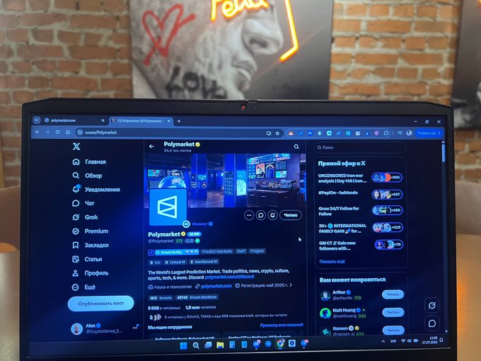 A laptop screen displaying the X (Twitter) profile of 'Polymarket' (@Polymarket), a prediction market platform for news, politics, crypto, culture, and more, shown in what appears to be a Russian-language interface with options like 'Главная' (Home) and 'Обзор' (Explore) visible in the sidebar.