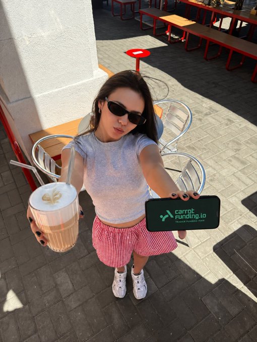 A woman wearing sunglasses, a gray t-shirt, and red gingham shorts holds a caramel drink with cream in one hand while taking a photo holding a phone with a 'carrot funding.io' green case, taken from a high angle in an outdoor café setting.