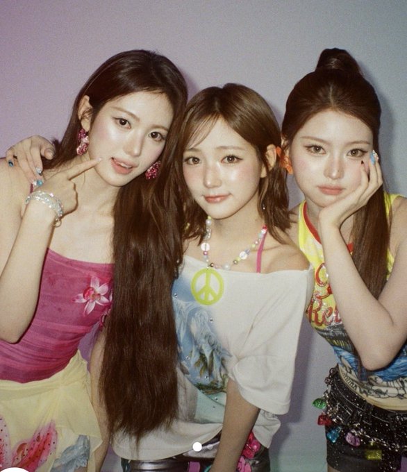Three young women pose closely together in a softly lit portrait, wearing colorful outfits with playful accessories including a neon peace-sign necklace and statement earrings.