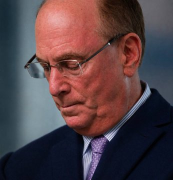 A close-up portrait of a balding older man with glasses, wearing a dark suit and a purple tie, looking downward with a serious expression.