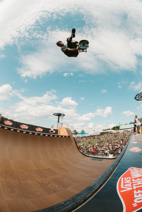A skateboarder performing an aerial trick high above a half-pipe ramp at what appears to be a Vans-sponsored skate event.