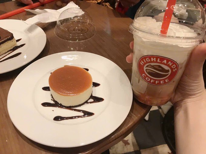 A hand holds a Highlands Coffee iced drink next to a plate featuring a flan caramel dessert decorated with chocolate sauce.