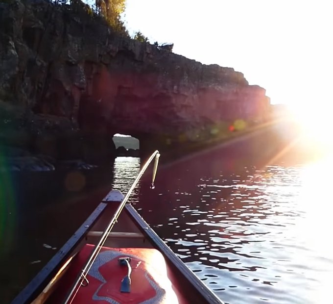 A view from a canoe approaching a cave opening in a rocky cliff, with bright sun glare reflecting off the water.