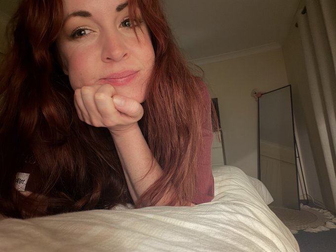 The image features a woman with long red hair lying down on her bed, smiling at the camera. She has her hand resting on her face as if posing for a photo. The background of the image shows a bedroom setting with a mirror hanging above the bed. There is also a pillow visible in the scene.