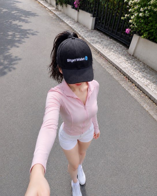 A selfie of a young woman wearing a black 'Bitget Wallet' cap, a pink zip-up long-sleeve top, and white shorts, standing on a street with a metal fence and flowering plants in the background.