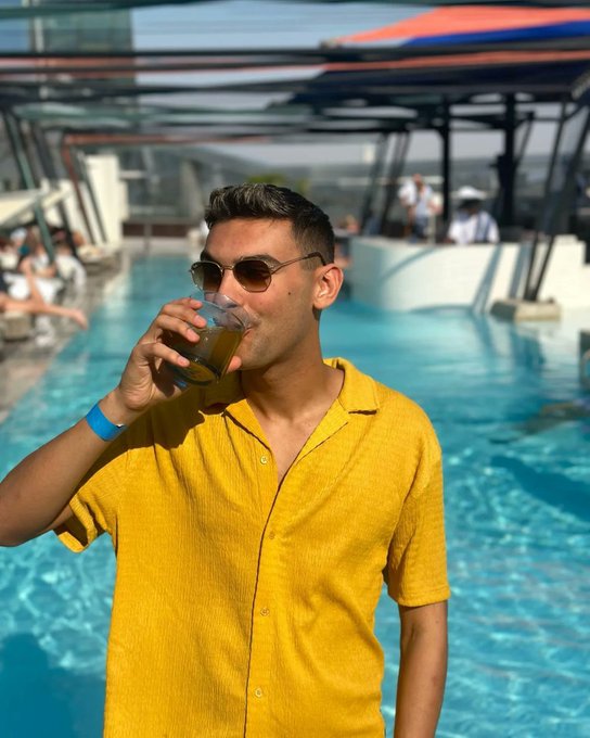 A man in a yellow short-sleeved shirt and sunglasses drinks from a glass while standing by a swimming pool with lounge chairs in the background.