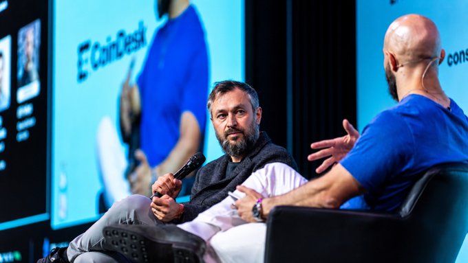 Two men having a conversation on stage at what appears to be the CoinDesk conference, with one bearded man holding a microphone while the other gestures while speaking.