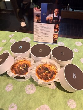 Photo of two open 'B' branded chocolate boxes revealing what appears to be a chocolate-covered pizza or dessert, displayed on a green patterned tablecloth with a product information card behind them.
