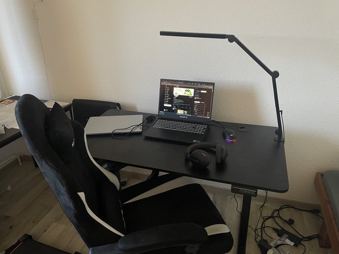 A home office setup features a black desk with a laptop, RGB gaming mouse, headphones, and a desk lamp, paired with a black and white gaming chair.