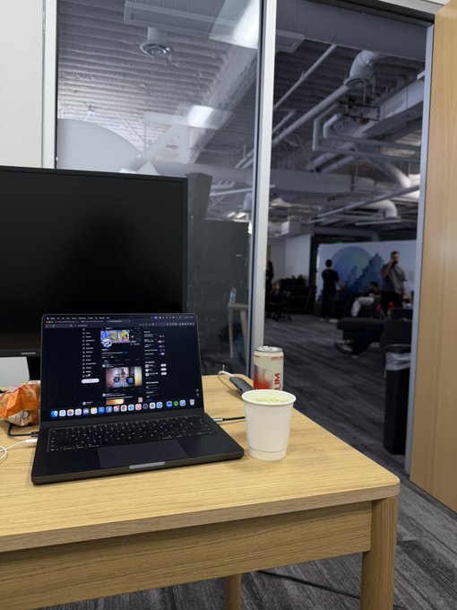 A laptop on a wooden desk displays a YouTube page, next to a paper cup and a soda can, with a view through a glass wall into an office space.