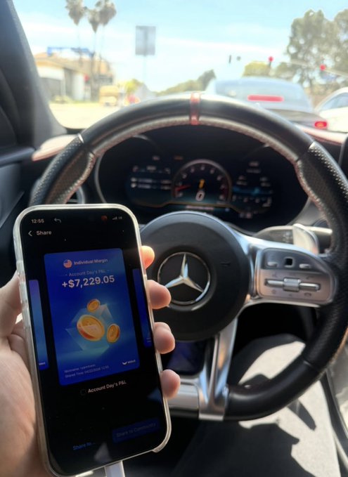 The image shows a person holding up their cell phone with a payment app, likely PayPal, displayed on the screen. They are also holding onto the steering wheel of a car while making the payment. The background includes other cars parked outside, indicating that they might be in an urban setting or at a parking lot.