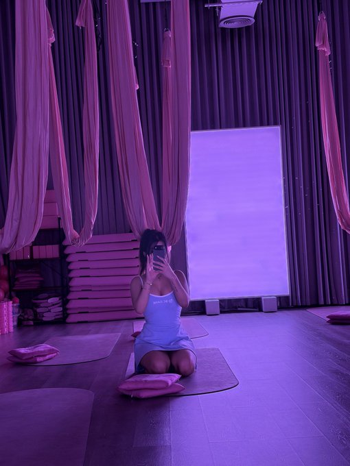 A person in a white tank top kneels on a yoga mat in a purple-lit studio with aerial silks, taking a mirror selfie.