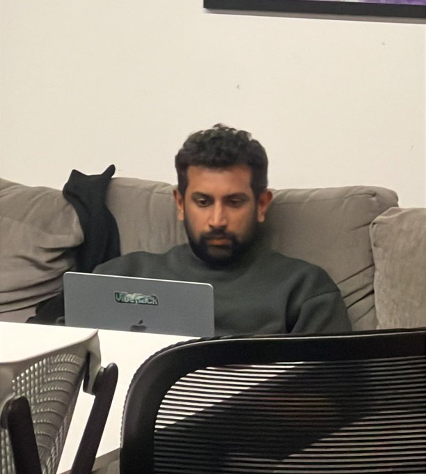 A man with a beard wearing a dark sweater sits on a gray couch working on a silver MacBook laptop with an "Underachievers" sticker. He appears focused on the screen in what looks like an office or casual workspace.