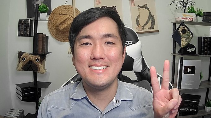 The image shows a smiling man holding up two fingers in a peace sign, sitting in front of shelves decorated with books, a pug pillow, and an "IN-N-OUT" sign. The post's playful tone and the casual, upbeat setting, along with the man's cheerful expression and gesture, reflect the kind of lighthearted, attention-grabbing content that can drive hype and speculation in stock or crypto communities.