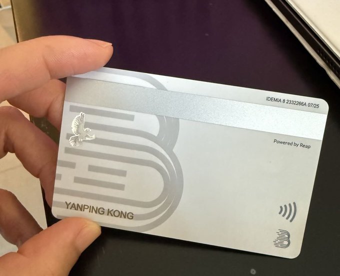 A hand holds a silver card labeled "YANPING KONG" with "Powered by Reap" and an IDEMIA serial number (8.2332268A 07/25), likely a Reap corporate card used as an alternative to U Cards for easily opening an account with just an ID card.