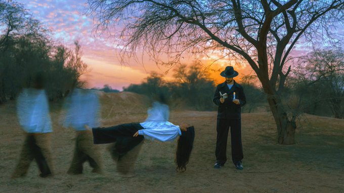 A long-exposure photograph at sunset showing blurred ghost-like figures walking, while one person stands still holding a white figure horizontally, and another wears a hat and holds sticks.