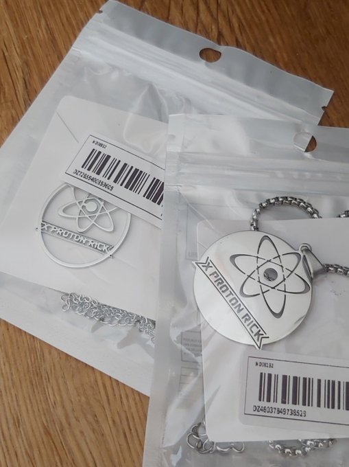 Two sealed plastic merchandise bags sit on a wooden surface, each containing a silver "XPROTONRICK" keychain/pendant with an atom logo and ball-chain, labeled with barcodes "X28612" (024037849738503) and "X28632" (024037849738529). The image showcases branded XPR Network/Proton Rick collectible merchandise being shipped, aligning with the promotional post highlighting the XProtonRick branding within the Metal Blockchain ecosystem.