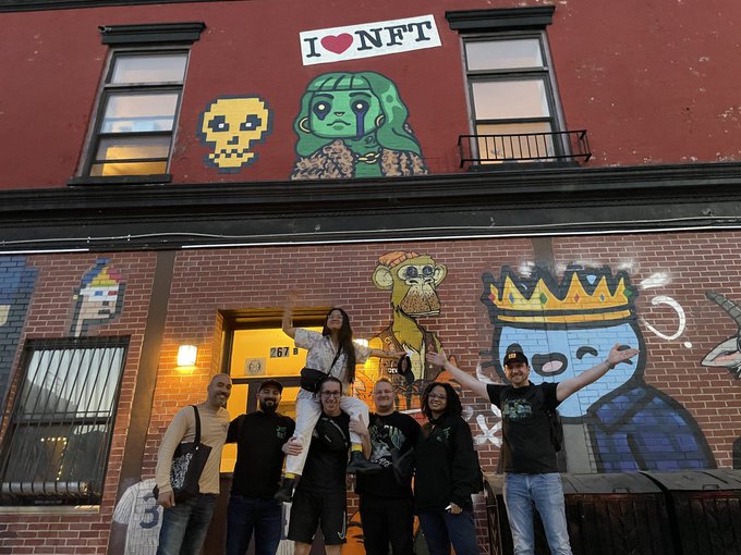 A group of people posing joyfully on a stoop in front of a brick building featuring an 'I ♥ NFT' mural and other crypto-themed artwork.