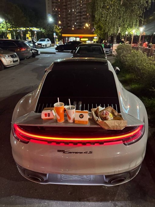 The rear view of a silver Porsche 911 Carrera 4S parked at night, with its spoiler raised and fast food drinks and bags sitting on the engine cover.