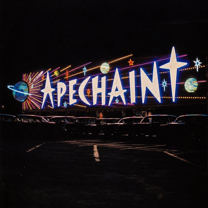 A nighttime photograph of a large, brightly illuminated sign that reads 'APECHAIN' in stylized white letters, surrounded by neon stars and planets, with cars parked in a lot in the foreground.