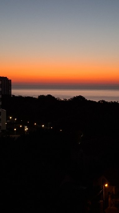 A serene sunset view over the ocean with a gradient sky of orange and deep blue, silhouetting dark tree-covered hills and streetlights in the foreground.