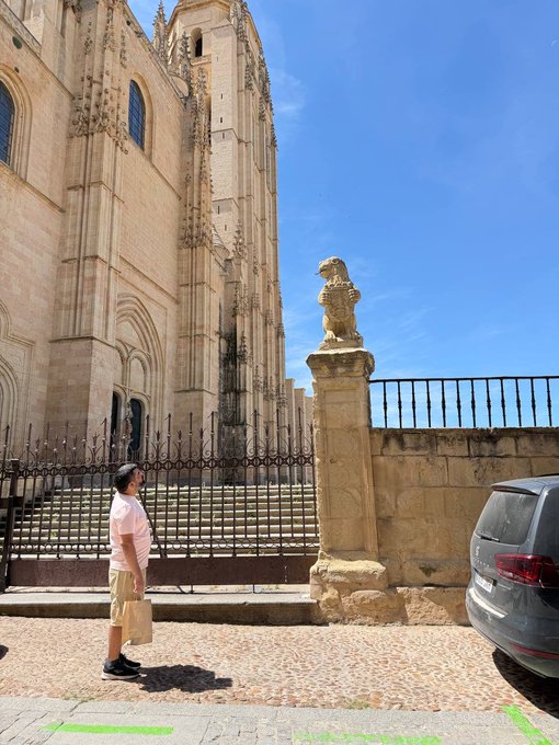 The image features a man standing next to a stone building with a statue of an eagle perched atop it. The man appears to be dressed casually, wearing shorts and a t-shirt. A car can also be seen parked nearby, adding context to the scene. The photo is taken from a low angle, making the man and the building appear larger than they actually are. The caption provided in the image seems to be related to an event or gathering taking place at the stone building, possibly involving the @solana Summit by @SuperteamDE.