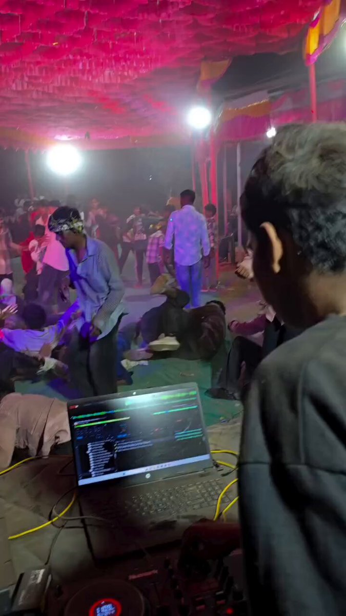 The image shows a DJ booth with a laptop computer sitting on top of it. The background reveals that there are people dancing around inside the tent-like structure, which appears to be an indoor event space or club. A man wearing a black shirt is standing in front of the DJ booth, possibly managing the music and equipment for the event.