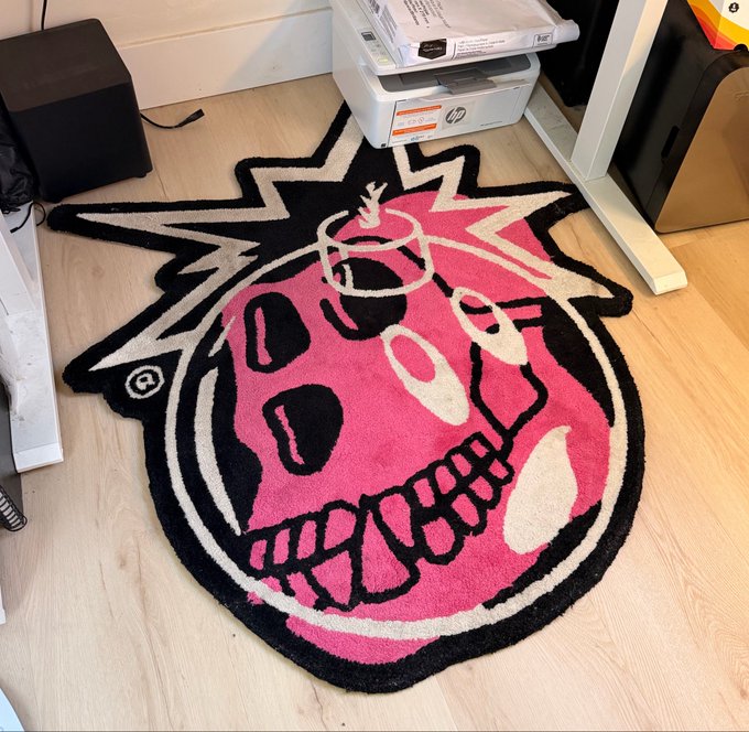 A pink and black shaggy rug featuring a stylized skull design with jagged lightning bolt accents, placed on a light wood floor near a printer.