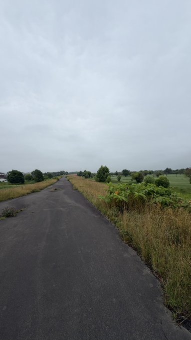 An empty, paved rural road stretches into the distance under a heavily overcast, humid sky, with tall grasses and trees lining the path. The image reflects the post's message about a humid morning running trip in Hokkaido, capturing the quiet, misty atmosphere before the run.