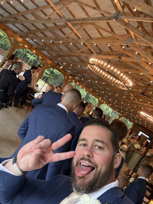 A bearded man in a navy suit takes a selfie at a wedding venue with a wooden beam ceiling, string lights, and other guests in formal wear in the background.