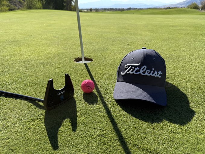 A pink golf ball near the hole on a golf course green, next to a putter head and a navy blue Titleist cap resting on the grass.