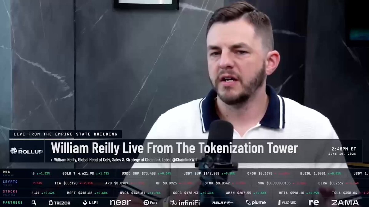 The image features a man wearing a white polo shirt with black stripes, standing behind a microphone while talking into it. The background of the image shows a gray wall with various logos displayed on it. The text "William Reily Live From The Bitcoinization Tower" is written in the image, and there are several other logos present as well. The man's position and the presence of logos suggest that he might be discussing or promoting something related to cryptocurrency or blockchain technology.