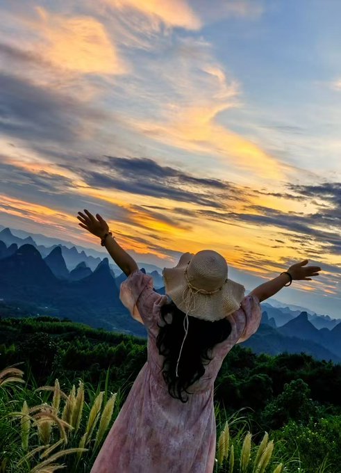A photograph of a woman in a floral dress and straw hat standing with her arms outstretched, facing a dramatic sunset over a mountainous landscape with lush greenery.