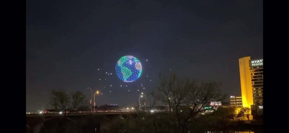 In the image, there are several colorful lights floating in the air above a city skyline at night. These lights appear to be coming from a large globe that has been illuminated with various colors. The globe is located near a bridge or overpass, which can be seen in the background of the photo. The post accompanying this image reads: "The largest Bitcoin symbol ever created by drones". This statement suggests that the globe's illumination might have some connection to Bitcoin or cryptocurrency, possibly as part of an event or celebration related to these digital assets.