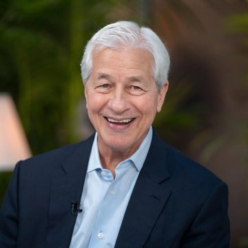 A smiling older man with silver hair, wearing a dark navy blazer over a light blue button-down shirt with a small lapel microphone, appears to be giving an interview against a softly blurred green backdrop. The image likely depicts a banking or financial leader discussing JPMorgan's expansion of JPM Coin to Ethereum, reflecting the post's theme of traditional banks adopting blockchain technology to enhance—though not replace—existing financial systems.