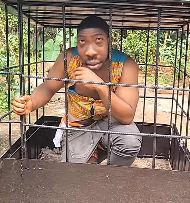 A man sits inside a metal cage outdoors, gripping the bars with his hands while wearing a colorful patterned tank top and gray shorts.